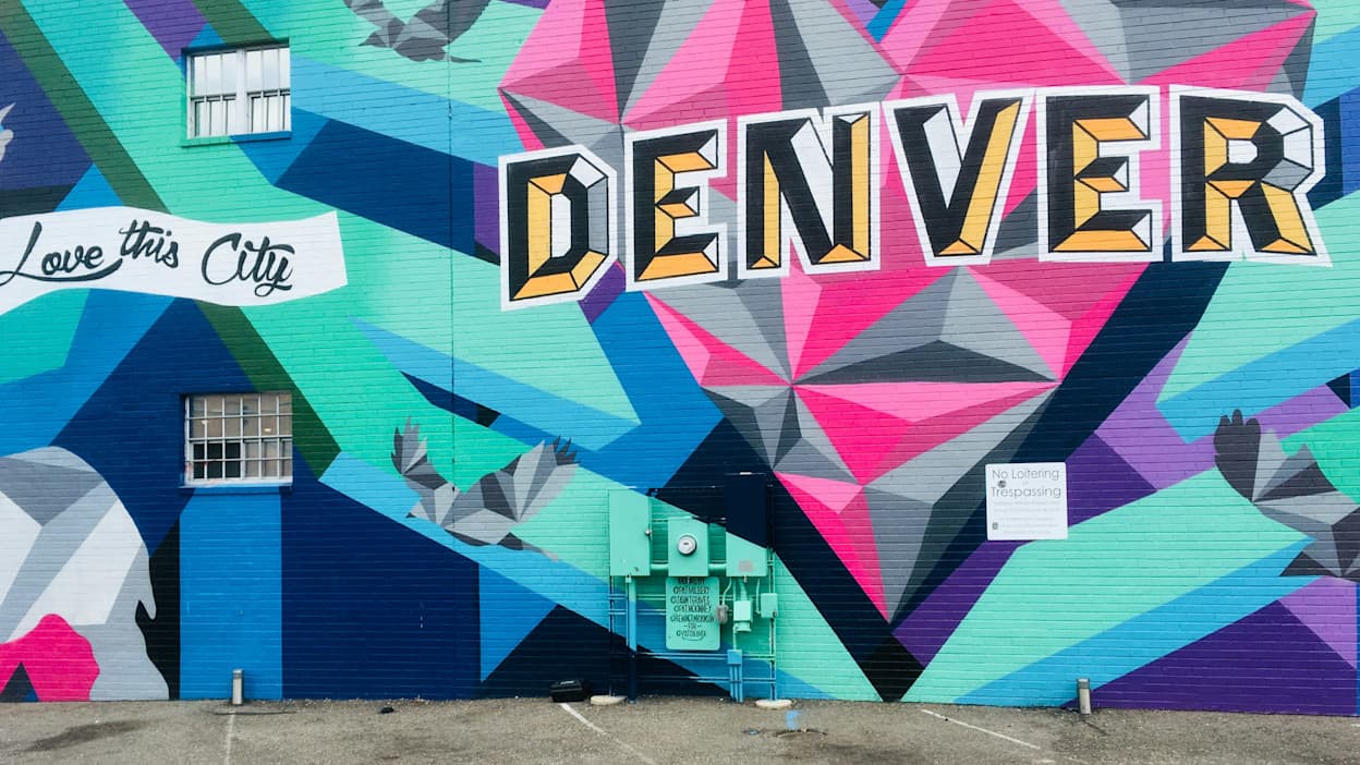 A mural that reads "Love this City DENVER" in black and yellow paint is painted on top of a turquoise, blue, pink, and purple geometric background. Taken in Denver, Colorado. 