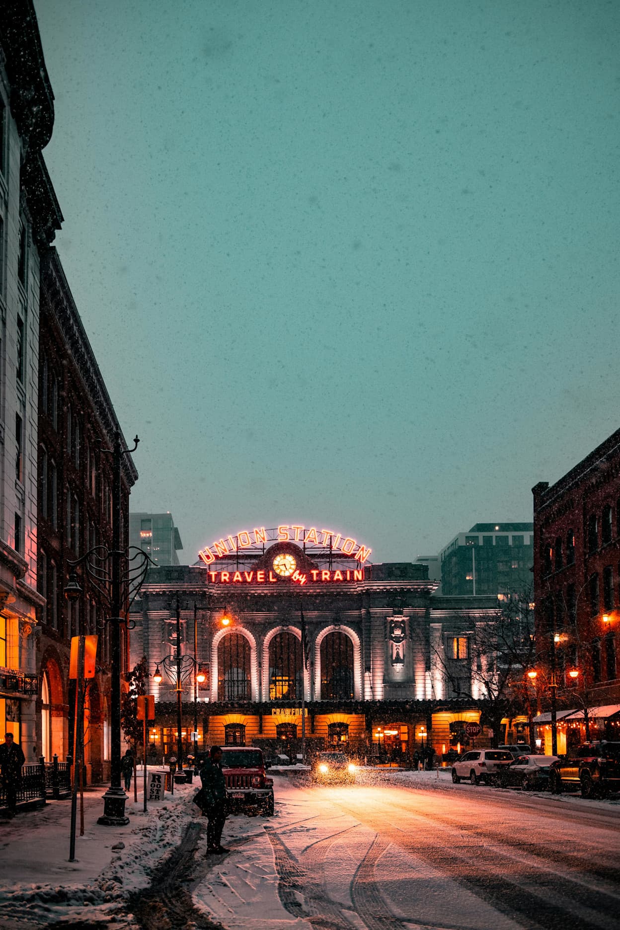 An image of the front of Union Station train station covered in snow in the LoDo neighborhood in Denver, Colorado. The lights are warm and some are casting a red hue on the image. 