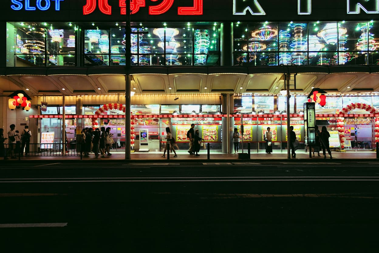 An image taken from the other side of the street of people standing on a sidewalk in front of a large retail storefront which is completely illuminated from within, and there are large letters on the outside partially cropped in the top of the image.