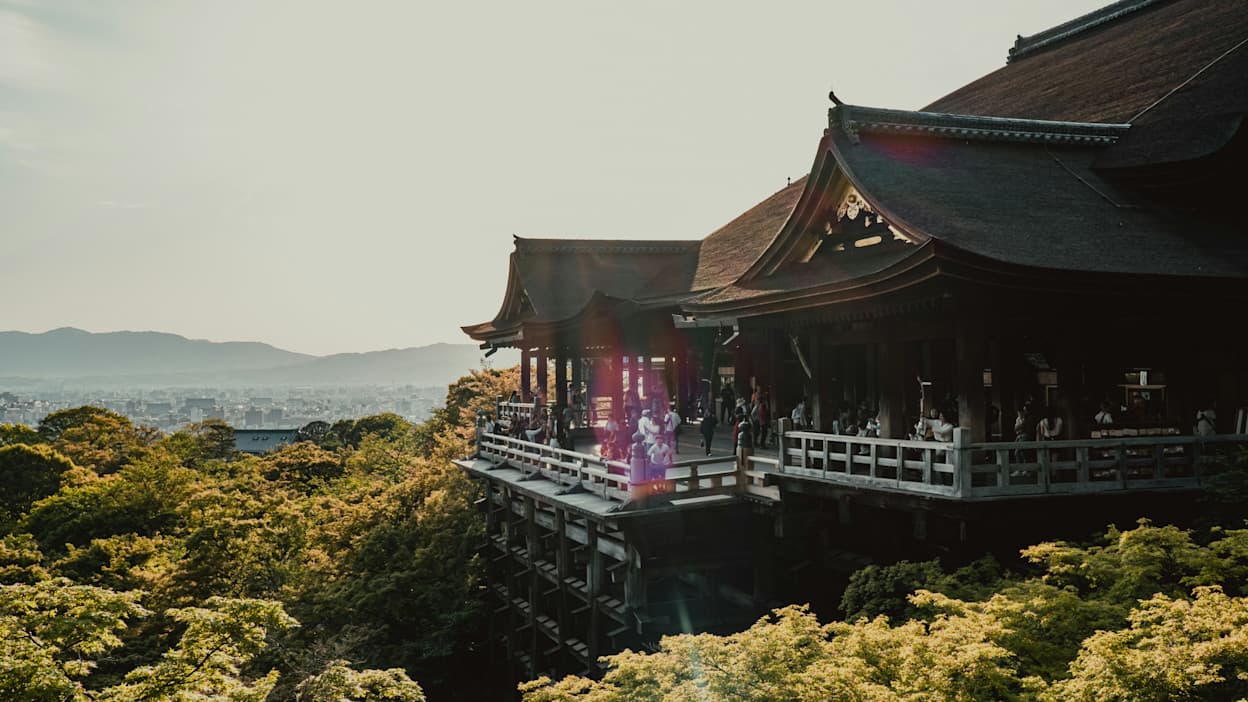 Taken from a hilltop in Higashiyama, an outdoor deck at a temple gazes across the Kyoto skyline. Mountains are visible on the horizon, and trees and greenery fill the foreground.