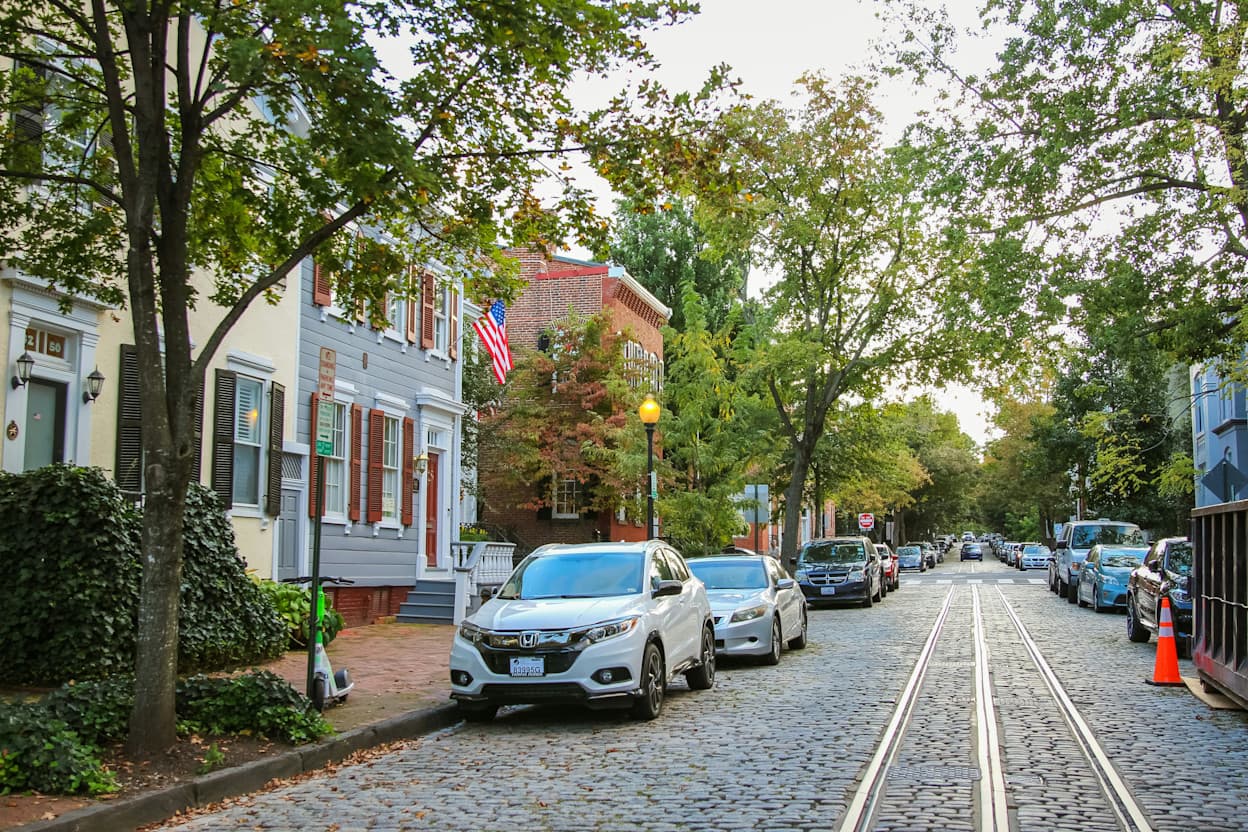Taken in the middle of a cobblestone road in Georgetown, Washington DC. Cars are parked along the road, and trees line the sidewalks. A train track runs through the center of the street, and houses line the road.