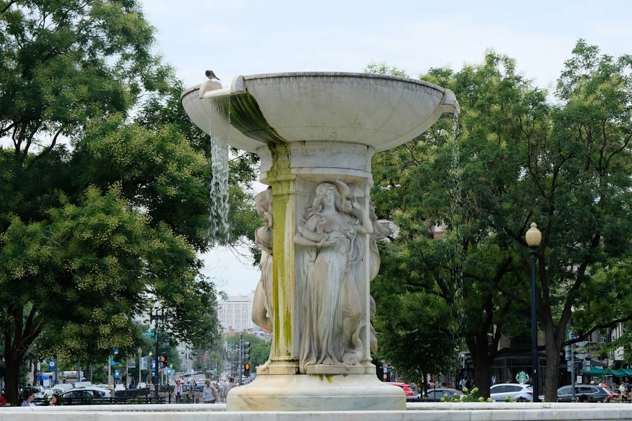 The famous fountain in the middle of Dupont Circle. Serving as the landmark of the Dupont Circle neighborhood, water falls from the bowl at the top of the fountain along the statues of women. Trees are in the background.
