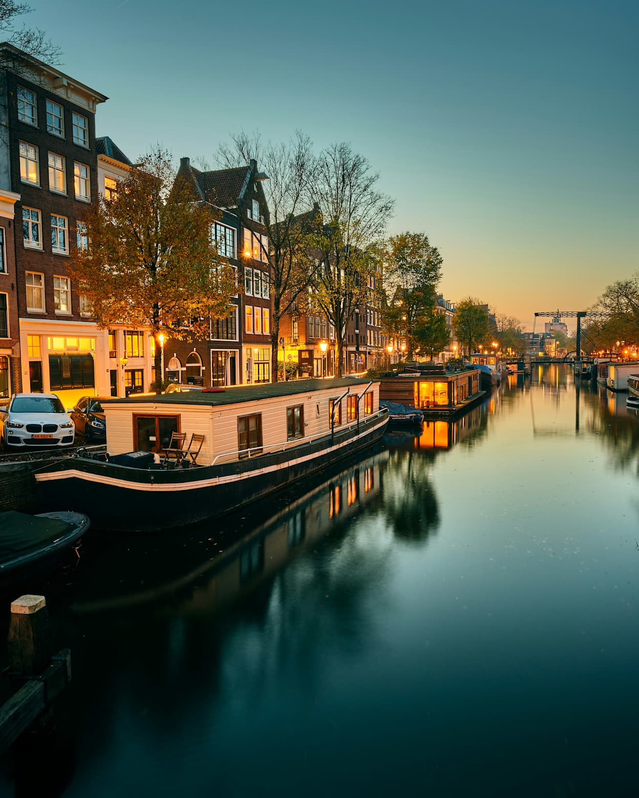 Taken along a canal in Amsterdam's Canal Belt neighborhood. Lined with houseboats, the canal is flat and reflects the boats and trees. Brick townhouses are in the background and are illuminated from within. 