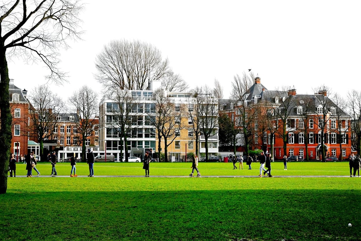 Taken in a park in the Museumkwartier of Amsterdam. The grass is a vibrant green, and many people are walking through the park. In the background, buildings line the road along with bare trees. 