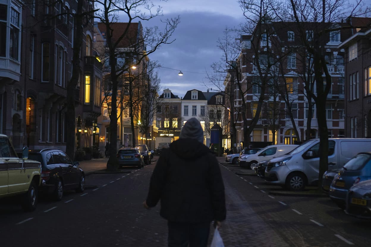 Taken along a cobblestone road in the De Pijp neighborhood in Amsterdam. Brick townhouses line the street, and the trees are bare in the wintertime. A man walks along the middle of the street between cars and towards more townhouses at the end of the road. 
