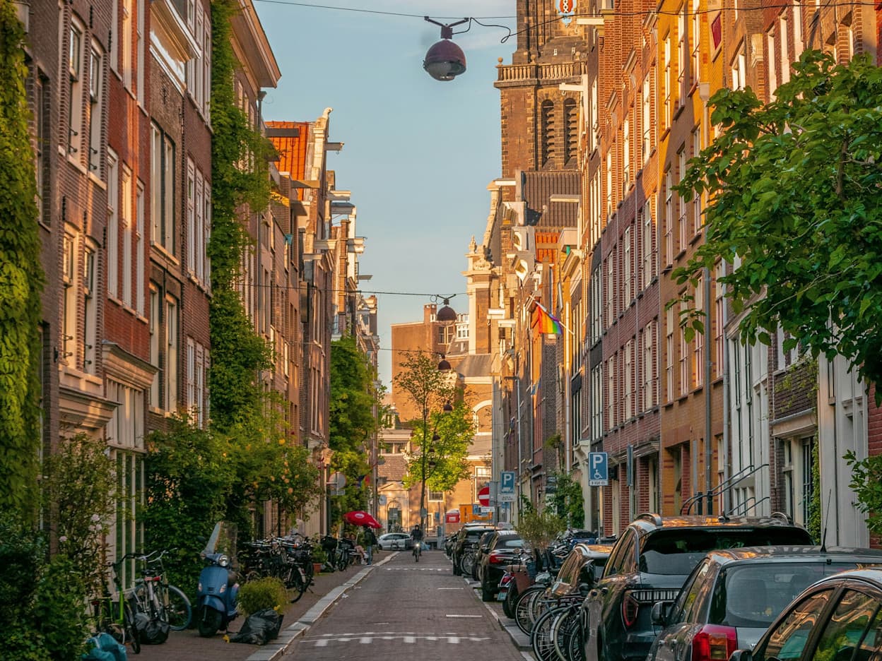 Taken along a cobblestone road in the Jordaan neighborhood in Amsterdam. Brick buildings and townhouses line the street, and a tall church protrudes in the background. Taken at sunset, a golden hue is cast on the buildings. 