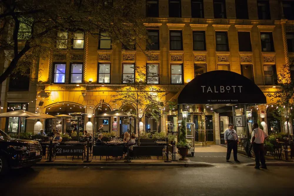 A street view image of the Talbott Hotel in the Gold Coast neighborhood of Chicago. The awning of the Talbott Hotel extends over the sidewalk, and lights are strung from the awning to cover the outdoor dining area along the sidewalk. People are standing in front of the hotel and are sitting within the dining area.