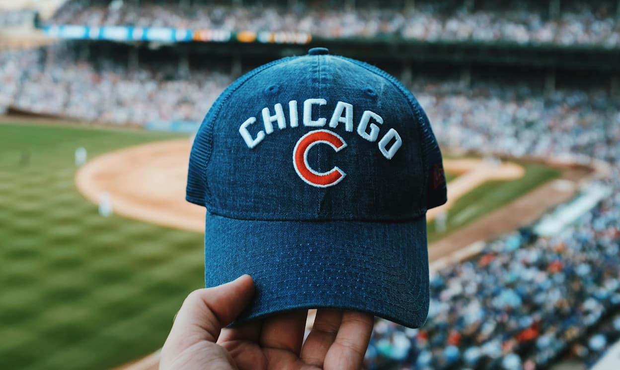 In focus is a hand holding a Chicago Cubs hat at Wrigley Field in Chicago. Beyond the baseball cap, out of focus, you can see a stadium filled with people at a mid-baseball game.
