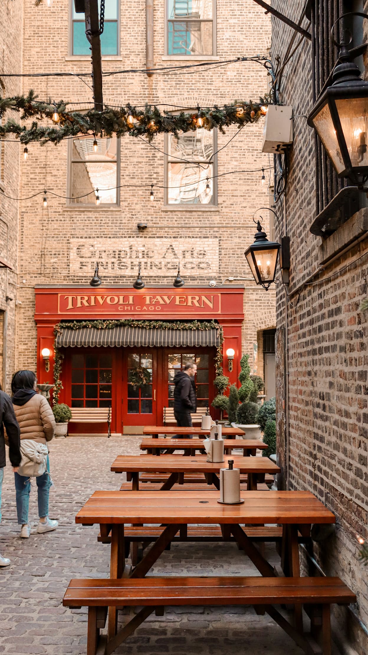 An image of Tivoli Tavern in the West Loop neighborhood in Chicago. Wooden picnic tables line the brick exterior in the alley, and vines with string lights are draped across the alley. A red storefront reads: "Tivoli Tavern"