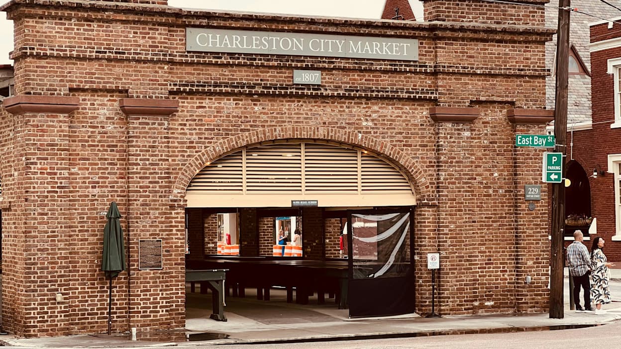 A street view image of the Charleston City Market in the French Quarter neighborhood in Charleston, South Carolina. The brick storefront has the store name on the front, and inside you can see tables and booths behind a barred entrance.
