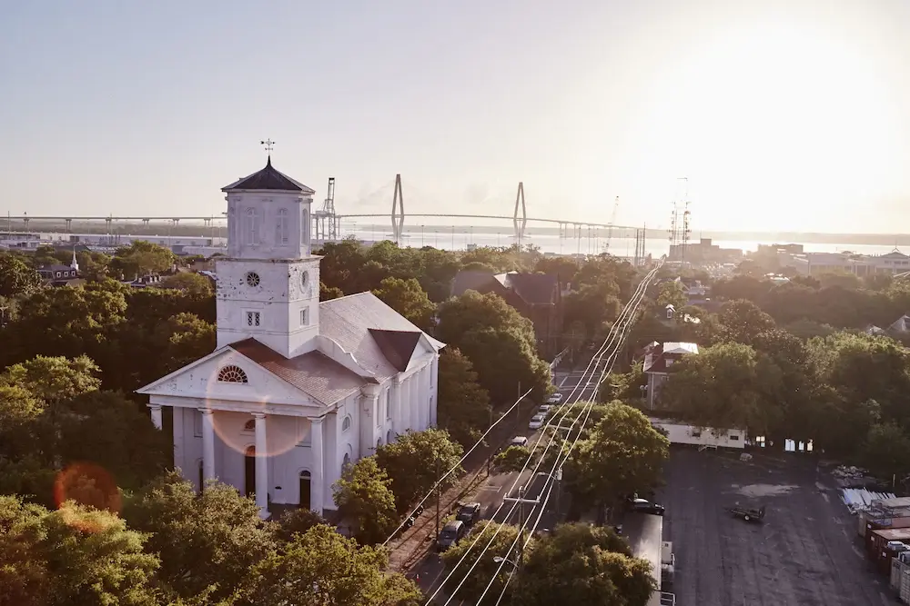 An image of a historic church in the Ansonborough neighborhood in Charleston, South Carolina. A bridge is seen in the background and lush trees fill the rest of the neighborhood.