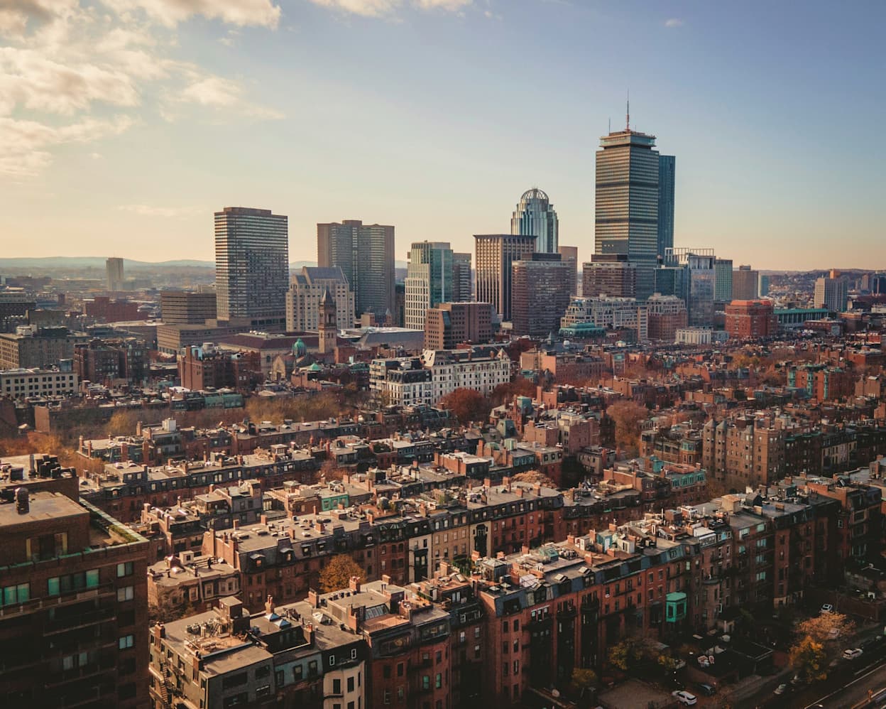 An aerial image taken of the uniform streets in the Back Bay neighborhood in Boston, MA. Beyond the brownstone residences, the taller buildings making up the Boston skyline protrude.