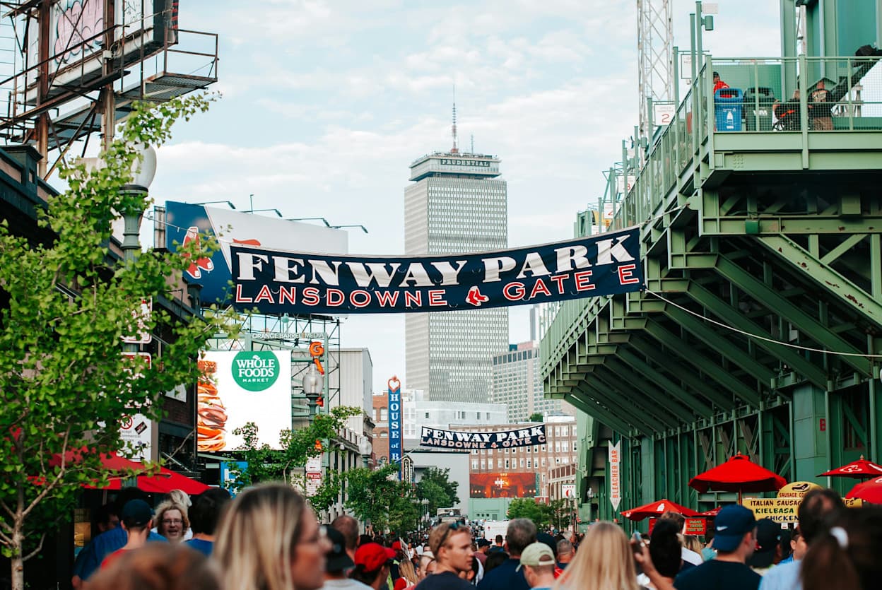 A banner across Landsdowne Street next to Fenway Park in the Fenway neighborhood in Boston, Massachusetts. People crowd the street in the foreground, and in the background the Prudential Center protrudes above the stadium and skyline.