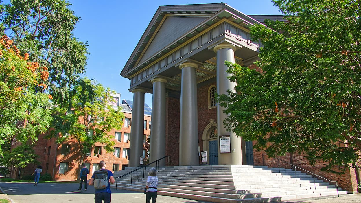 Taken outside one of Harvard University's signature brick and grandiose buildings with marble steps and details. Students walk outside the building, and lush green trees fill the space. Harvard University is in Cambridge, Massachusetts.