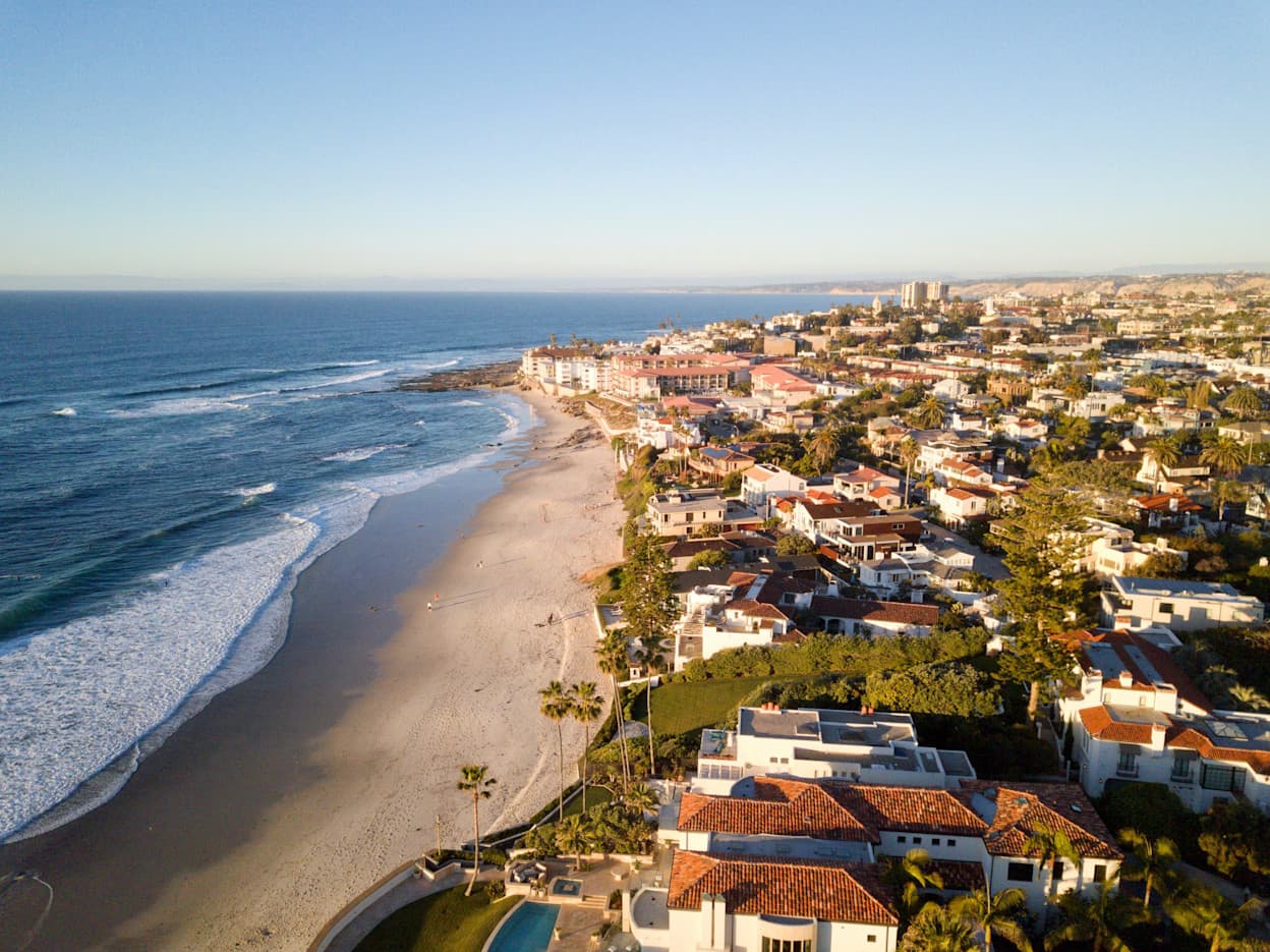 An aerial image of La Jolla, San Diego. The image captures large houses along the coastline, bordering on the white sandy beach and the low waves of the Pacific Ocean. Houses have terracotta roofs and there is a lot of greenery in the neighborhood.