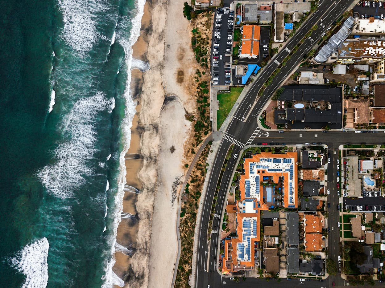 A bird's eye image of Carlsbad, California, where the ocean and beach comprise 1/3 of the image and the town along the beach the other. There are white caps from the waves rolling in along the white sand beach, and the homes and buildings along the beach are split by a road going through town.