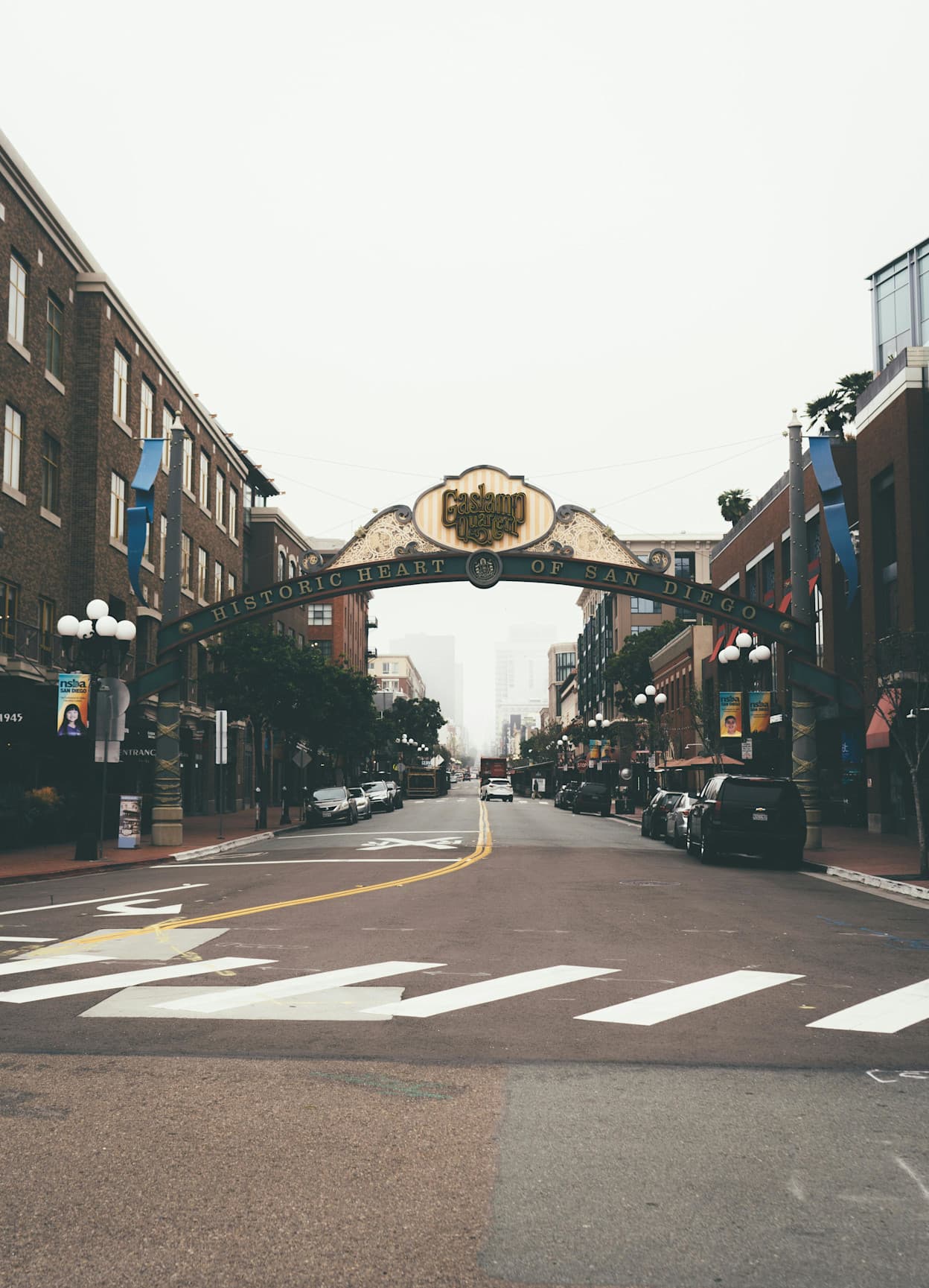 An image taken from a street in the Gaslamp Quarter neighborhood in San Diego. The image captures a sign acting as an entry to the neighborhood, which has "Gaslamp Quarter: Historic Heart of San Diego" written across it. Brick buildings line the street.