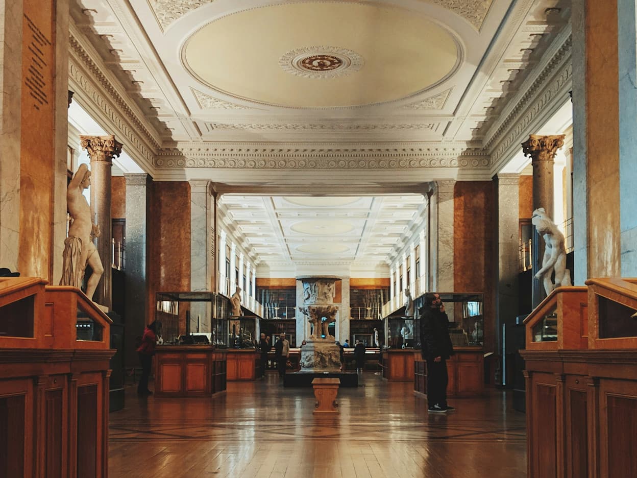 A reading room in the British Museum, in the Bloomsbury neighborhood of London. The reading room has solid wood floors and finishings, adorned with an ornate ceiling pattern. Old statues line the hallway walls, leading into the reading room.