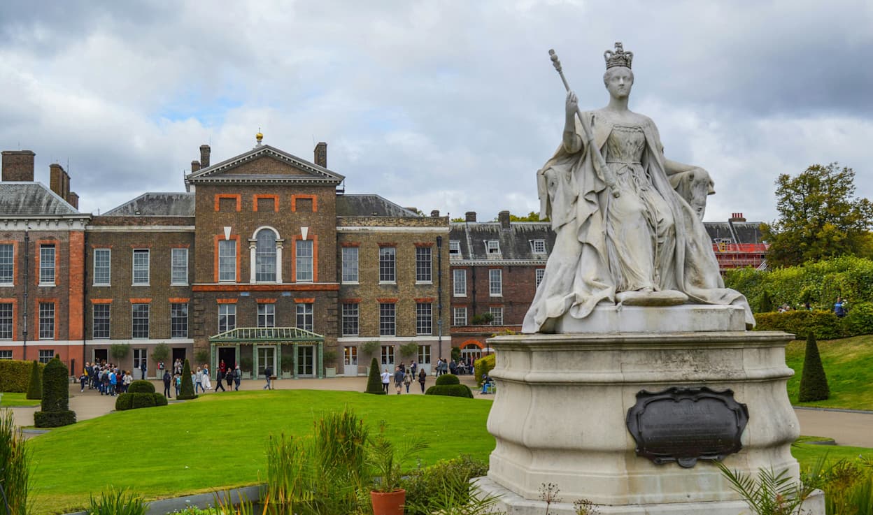 The brick exterior of Buckingham Palace with Queen Victoria Statue in London, a green lawn is between the two historical structures