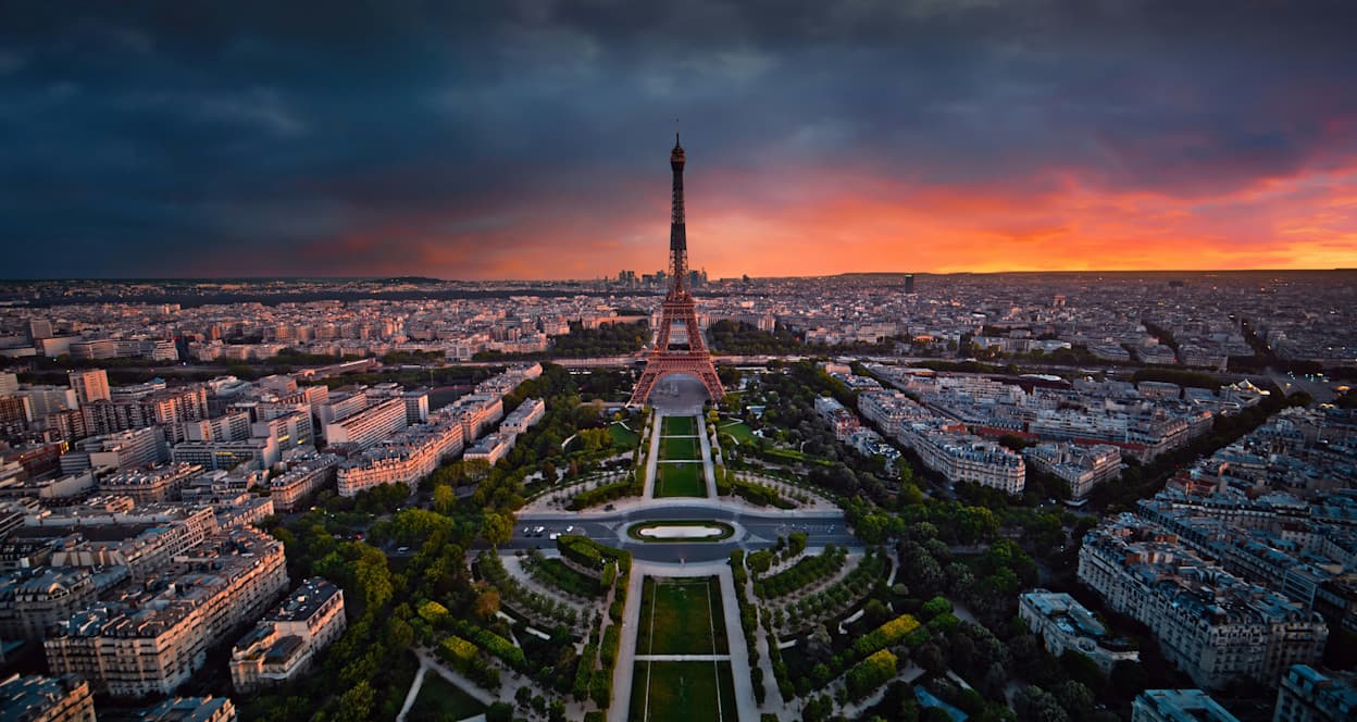 The Eiffel Tower protrudes above the green Champ de Mars park, surrounded by buildings. Taken at sunset, the sky is mostly dark with orange and pink clouds, casting an orange glow on the Parisian skyline. 