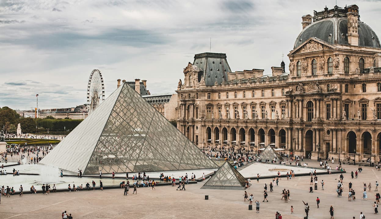 People walk around the glass pyramid in the courtyard of the Louvre, with the grand exterior of the Louvre museum behind it. In the background, a Ferris wheel peaks above the Louvre. 