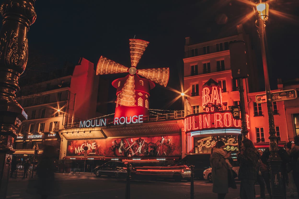 The historic Moulin Rouge building in Pigalle, Paris, is covered in red lights, some of which spell "Moulin Rouge" and on top of the structure, a large light-up windmill is also on display. 