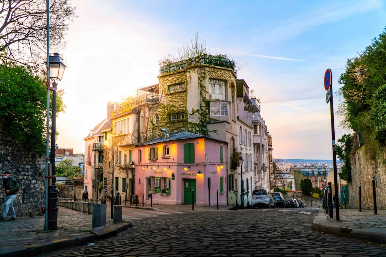 A historic corner building in the Montmartre neighborhood of Paris. The building is painted partially pink and the higher stories are covered in vines. The cobblestone road continues on in either direction, providing panoramic views of Paris. 