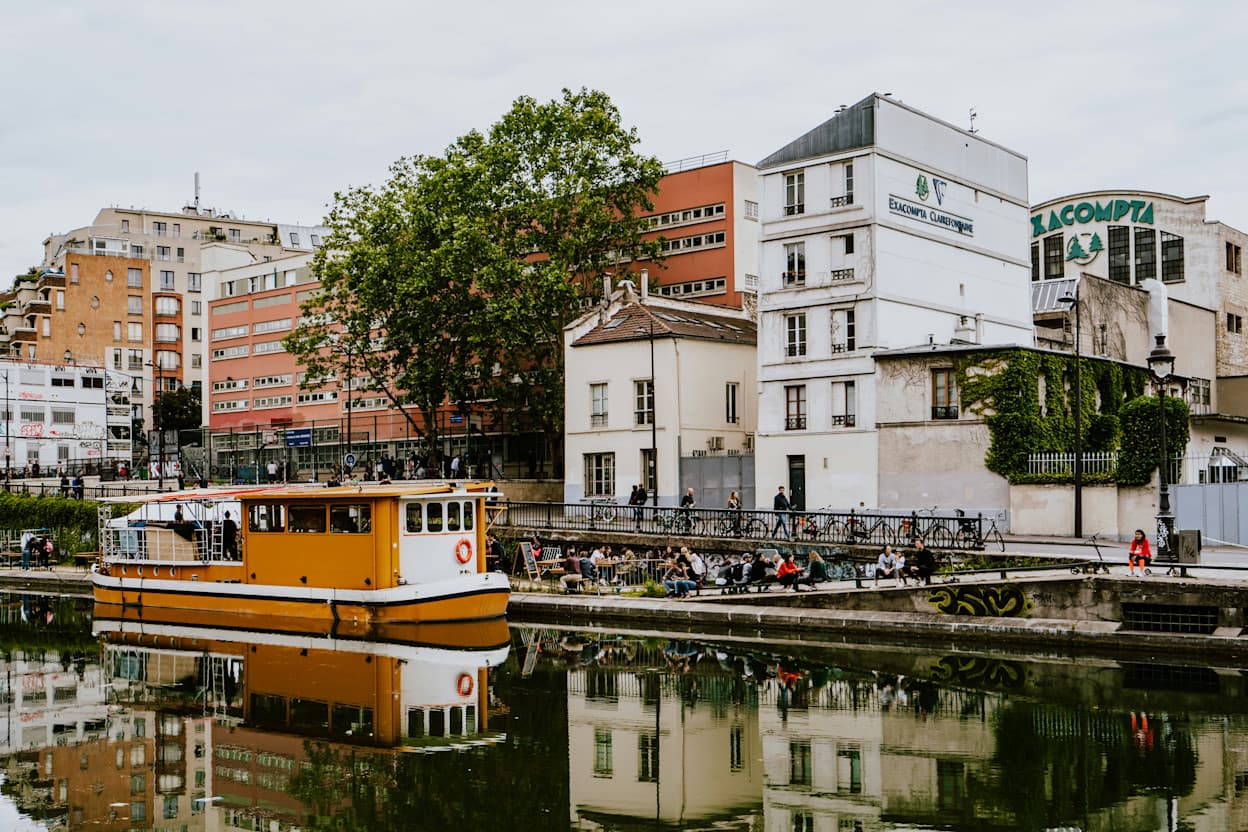 An orange boat is docked next to people sitting on a pier along Canal Saint-Martin, Paris. Buildings and trees line the canal. 