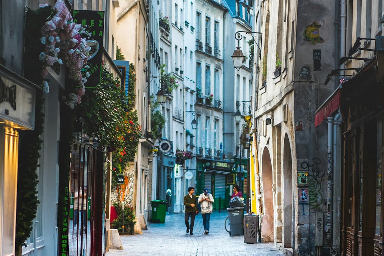 People walking through a narrow street in the Le Marais District of Paris. The buildings lining the road have stone embellishments, flowers and plants hanging from windows, and are painted different pastel colors. 