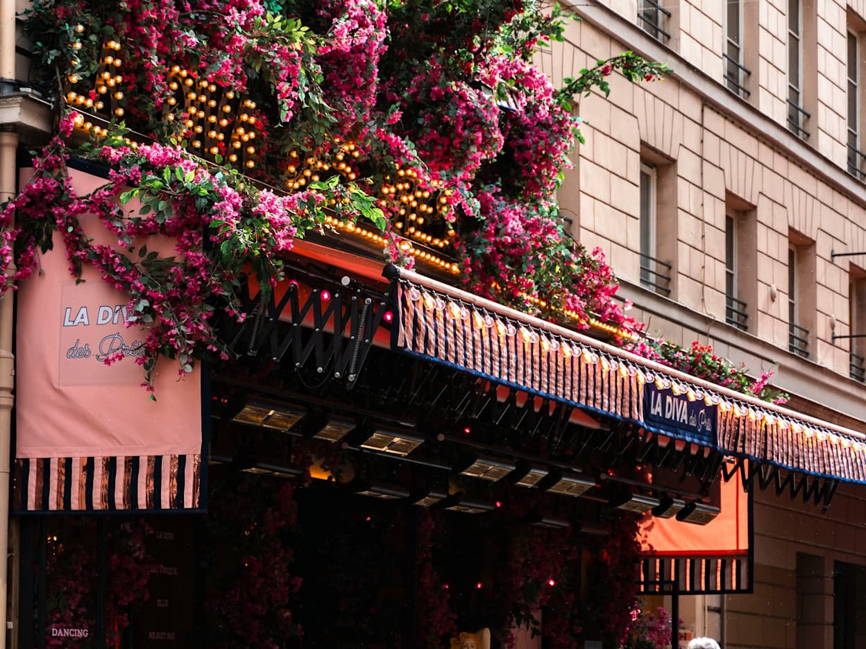 Pink bougainvillea flowers and greenery, along with a marquee-style sign with the restaurant's name, "La Diva des Pres," adorn the exterior awning of a restaurant in the Latin Quarter of Paris. 