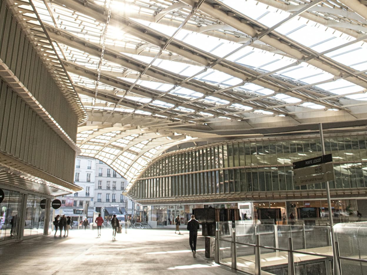 The modern Les Halles Shopping Center has a glass roof and glass walls supported by metal beams. People walk through the shopping center, which opens onto a street. 