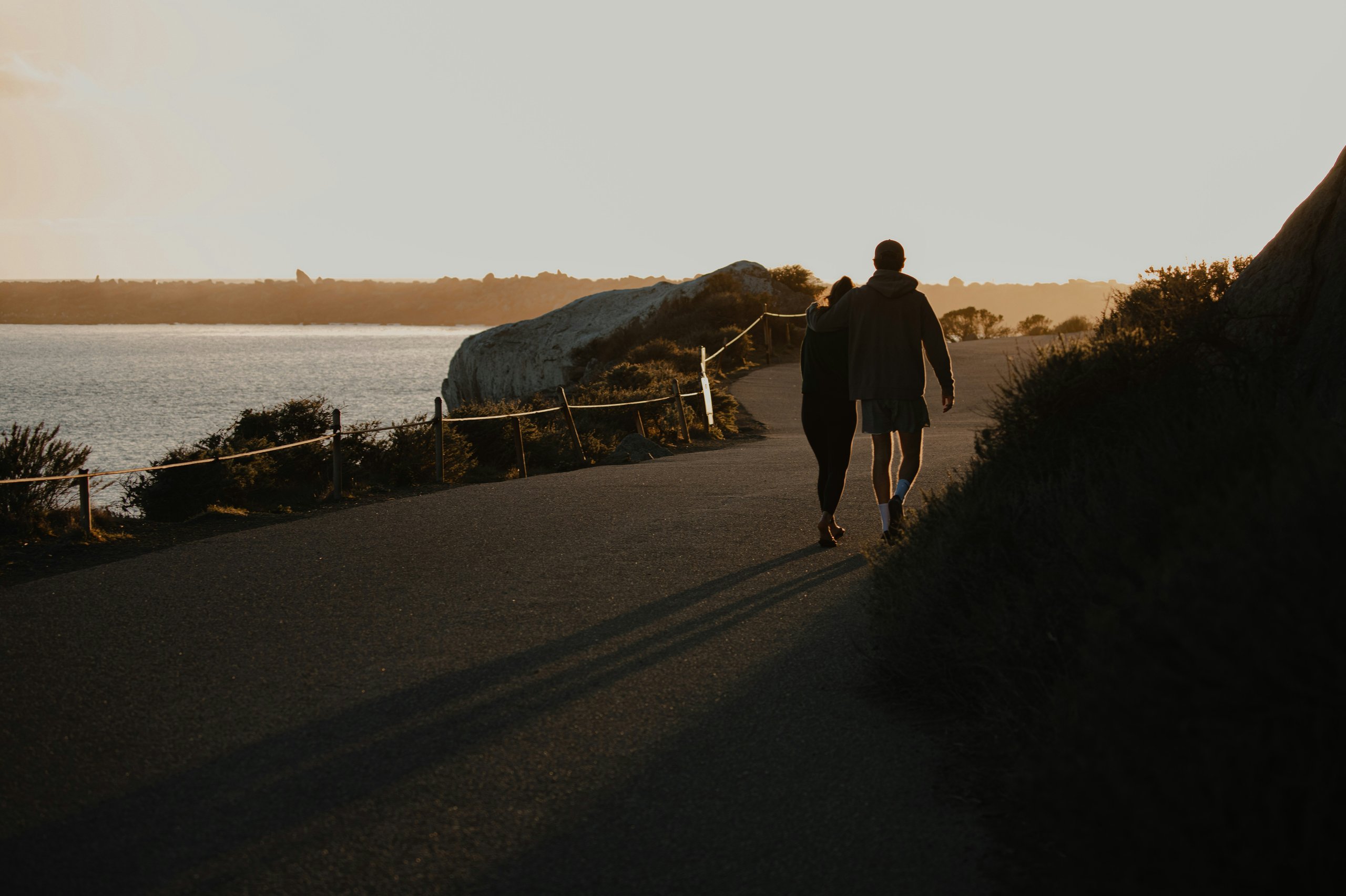 Couple walking arm-in-arm on a coastal path at sunset with long shadows stretching behind them.