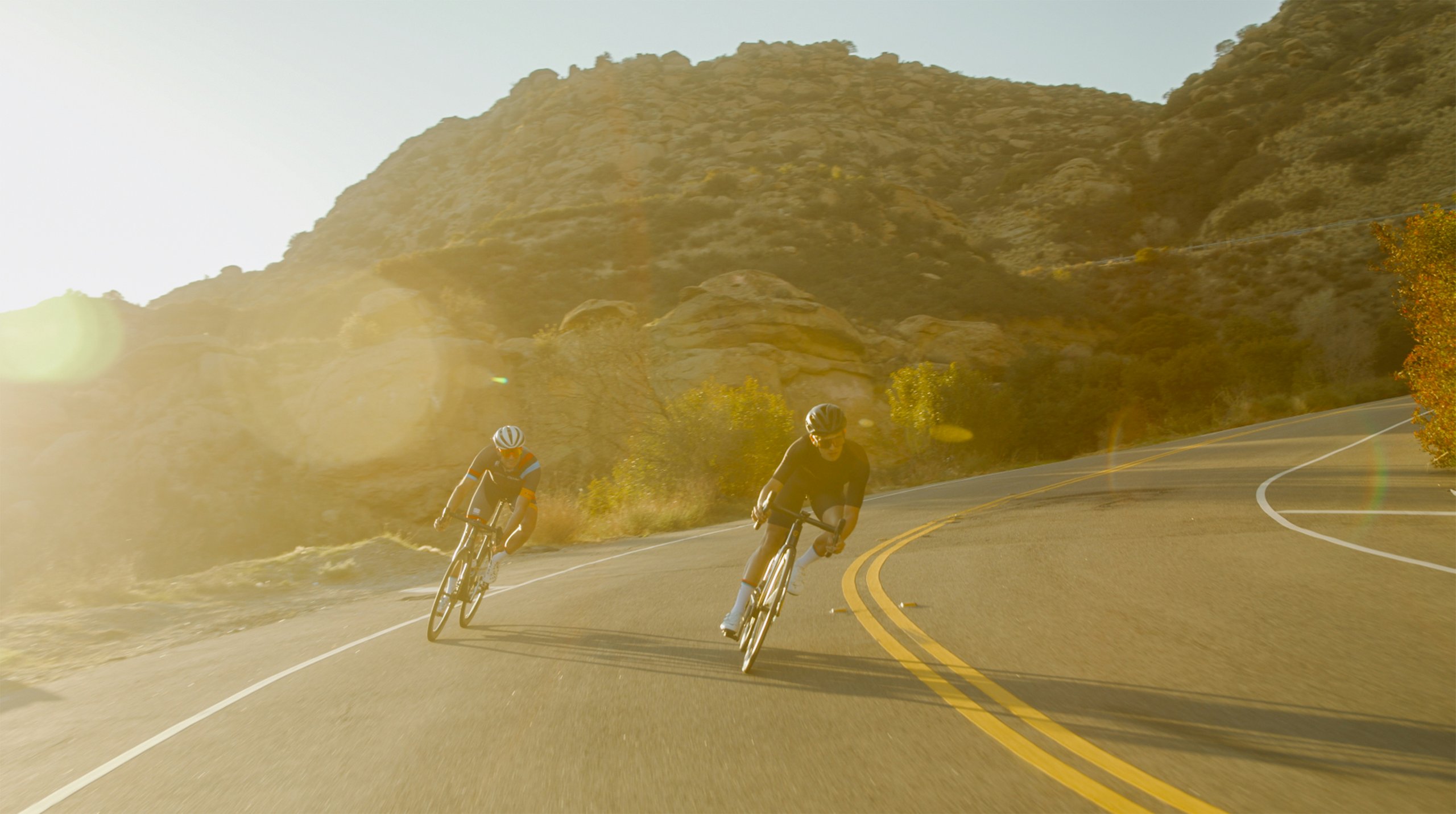 Two cyclists wearing helmets racing on a winding road near rocky hills with sunlight glare.