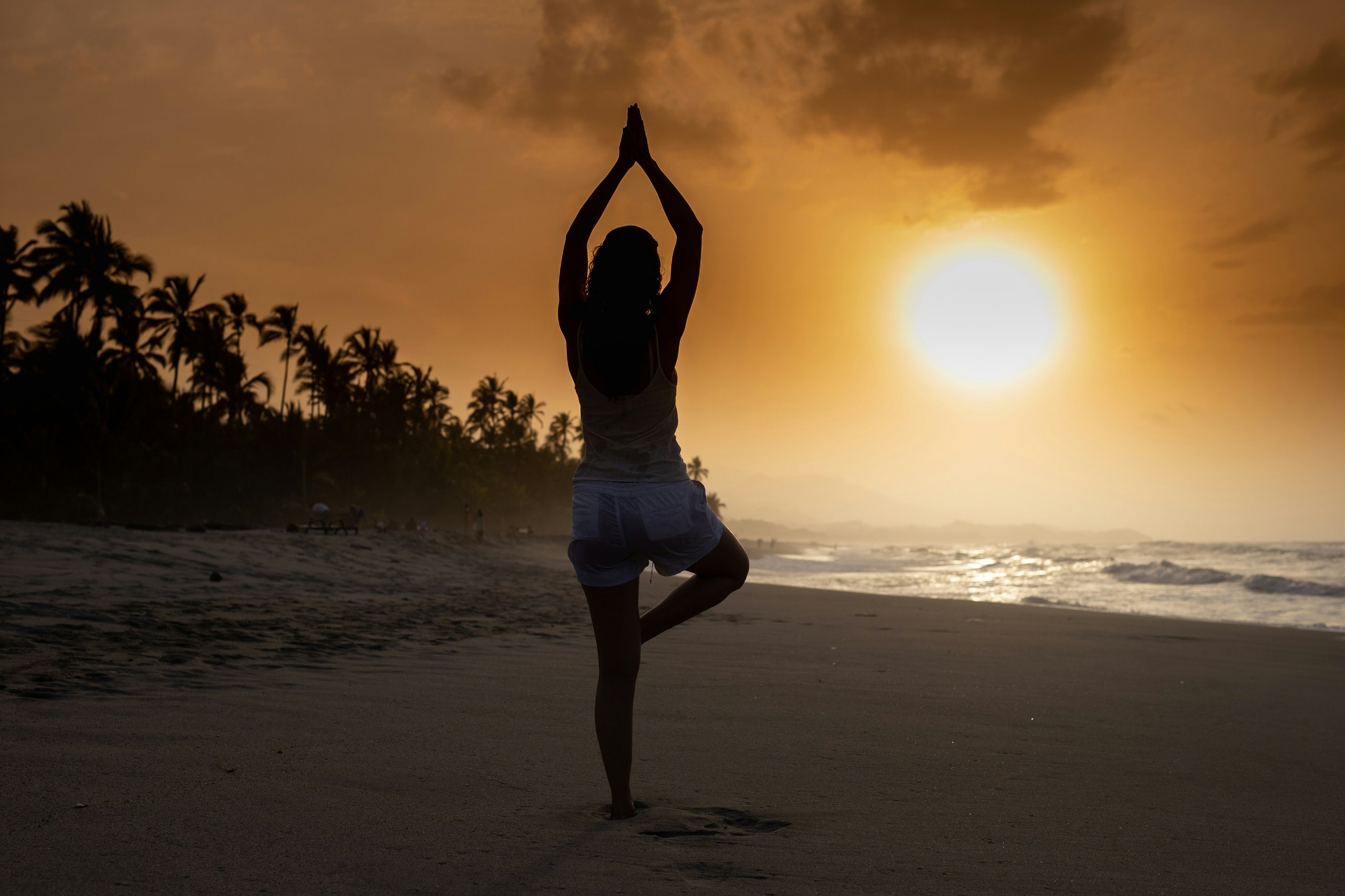 Person practicing yoga in tree pose on a beach at sunset with palm trees and ocean waves in the background.