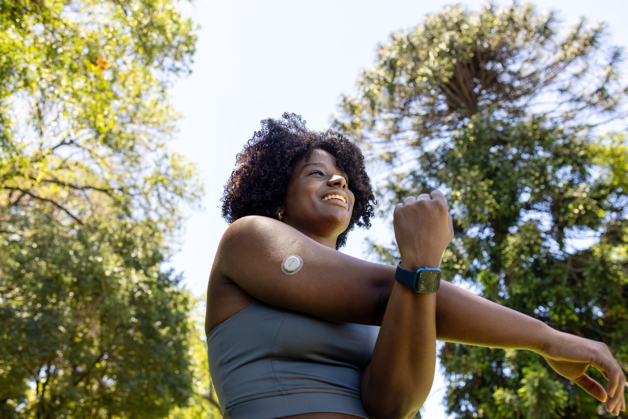Smiling woman outdoors stretching her arm wearing a fitness tracker smartwatch and a glucose monitor on her upper arm.