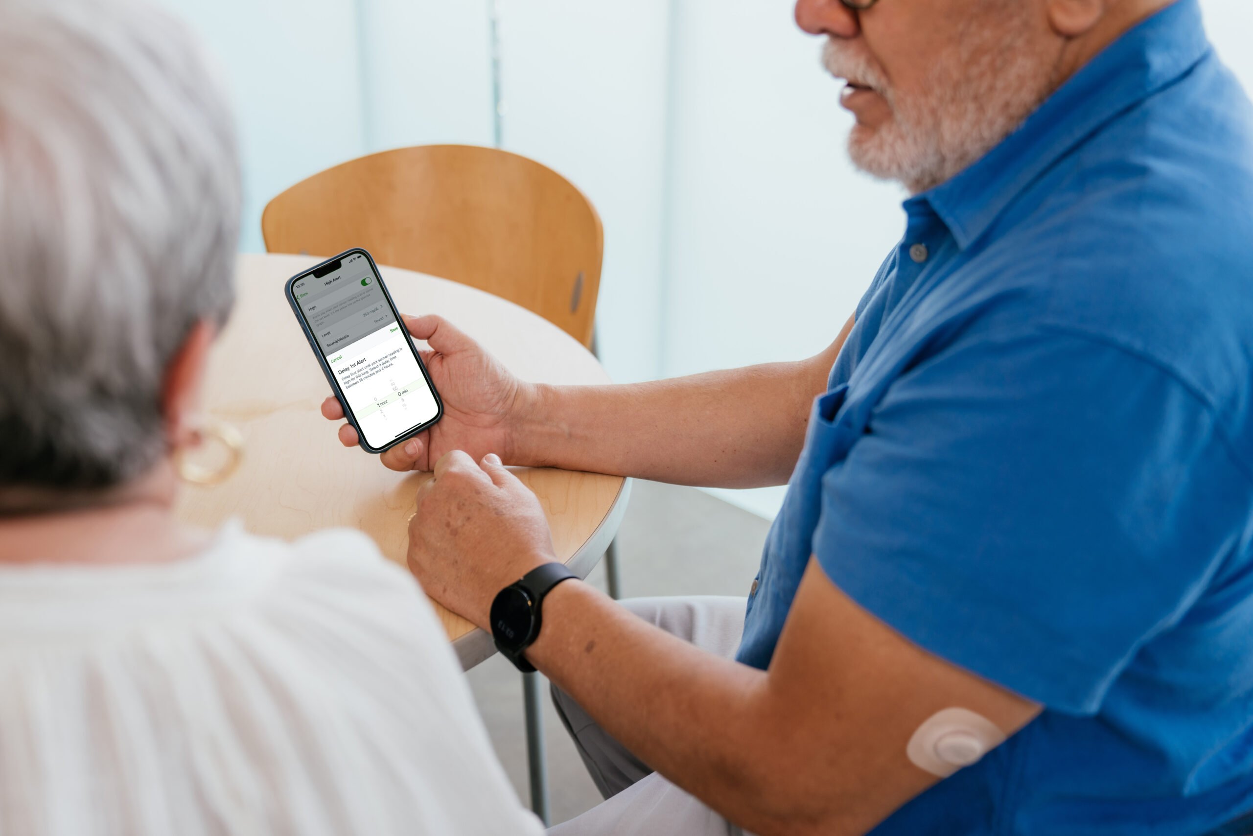 A man showing a woman a smartphone with a diabetes app displaying a glucose alert setup screen.