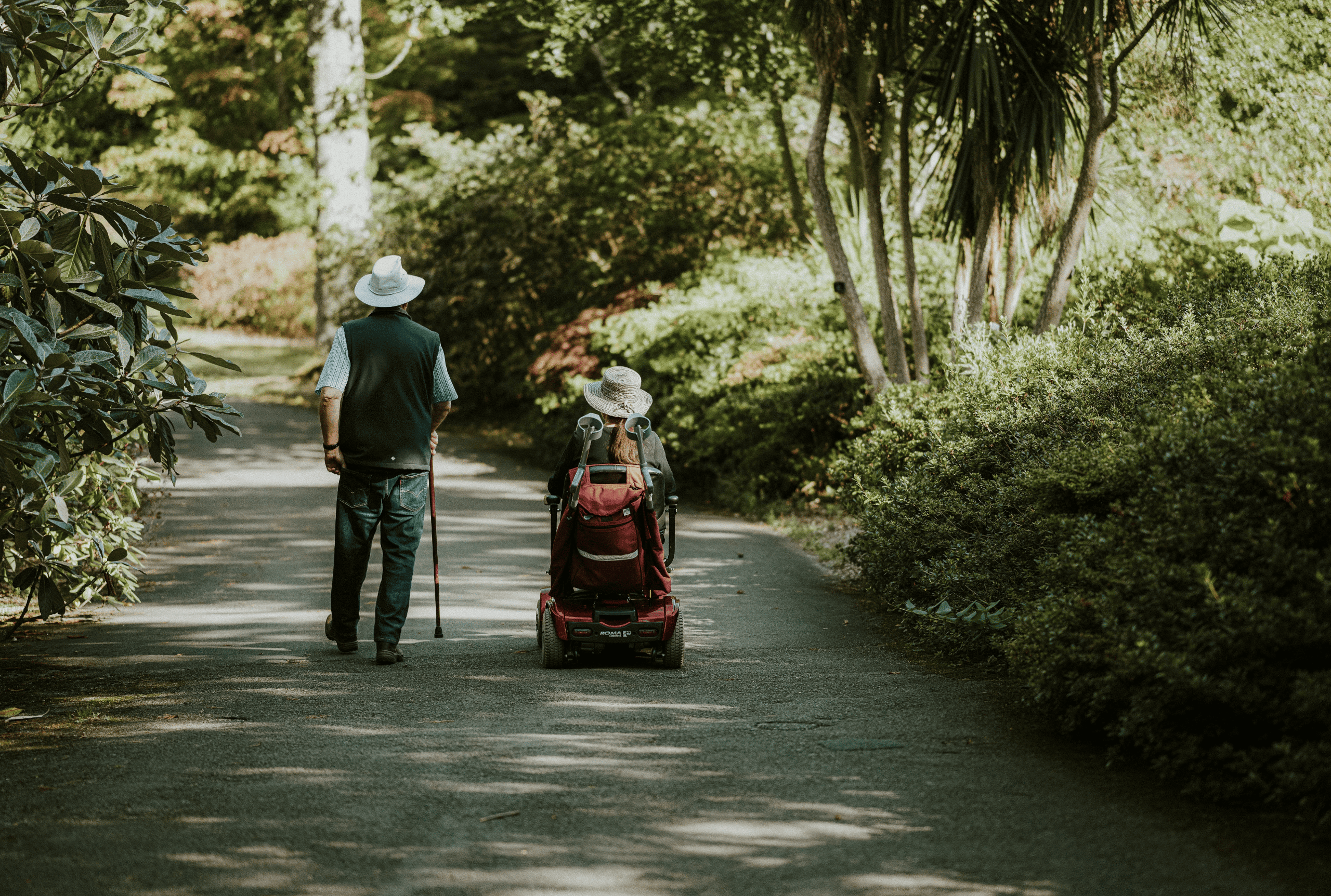 Man with cane walking beside a woman in a mobility scooter on a tree-lined paved path.