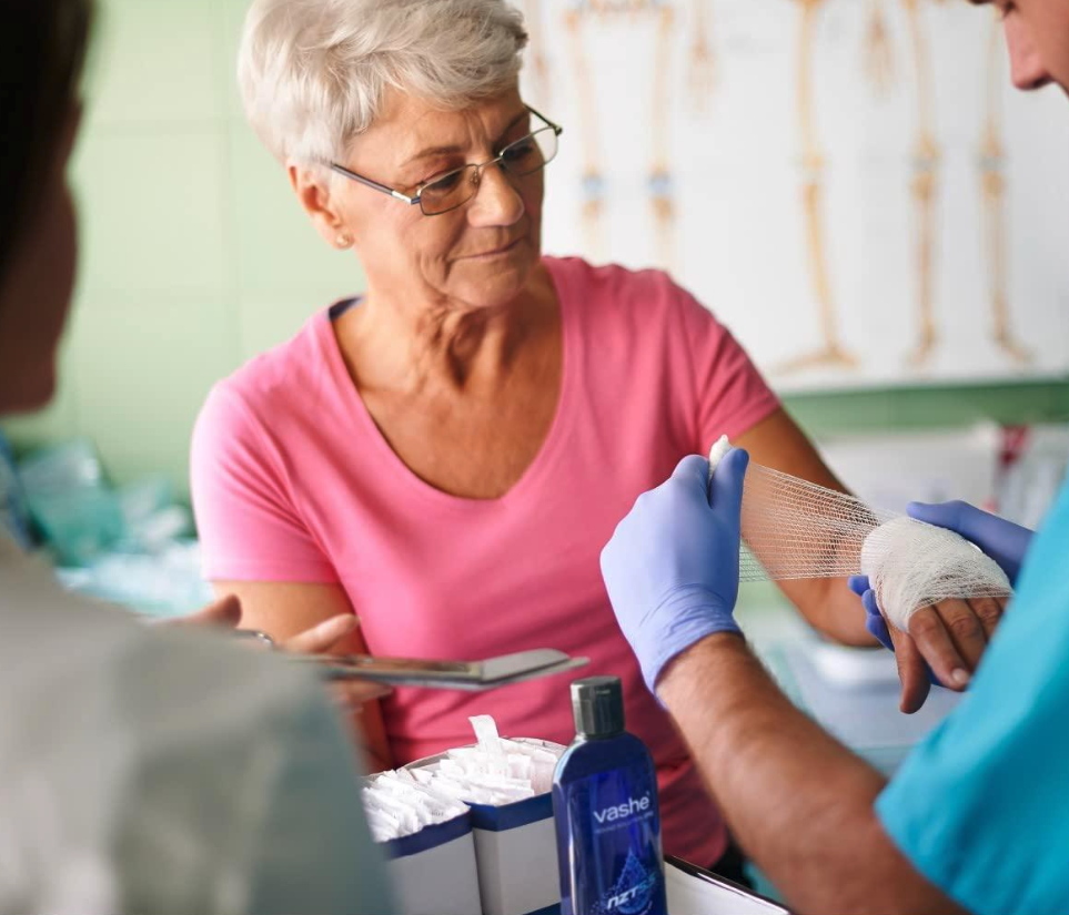 An elderly woman wearing a pink shirt receiving bandage care from a healthcare professional wearing blue gloves.