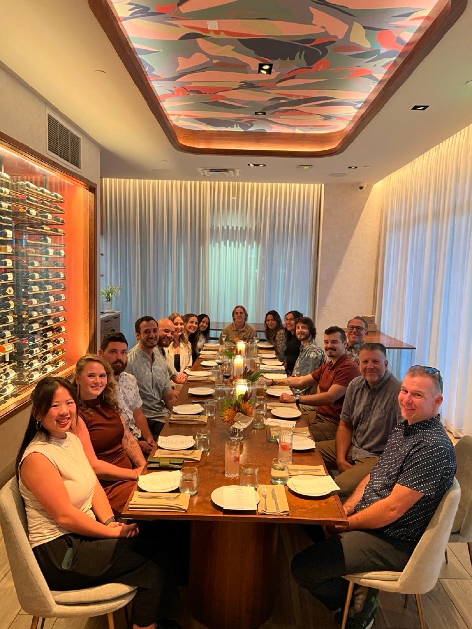 Group of people from CADE Medical team seated around a long dining table in a modern, warmly lit room with a colorful ceiling panel and a wine rack on the side.