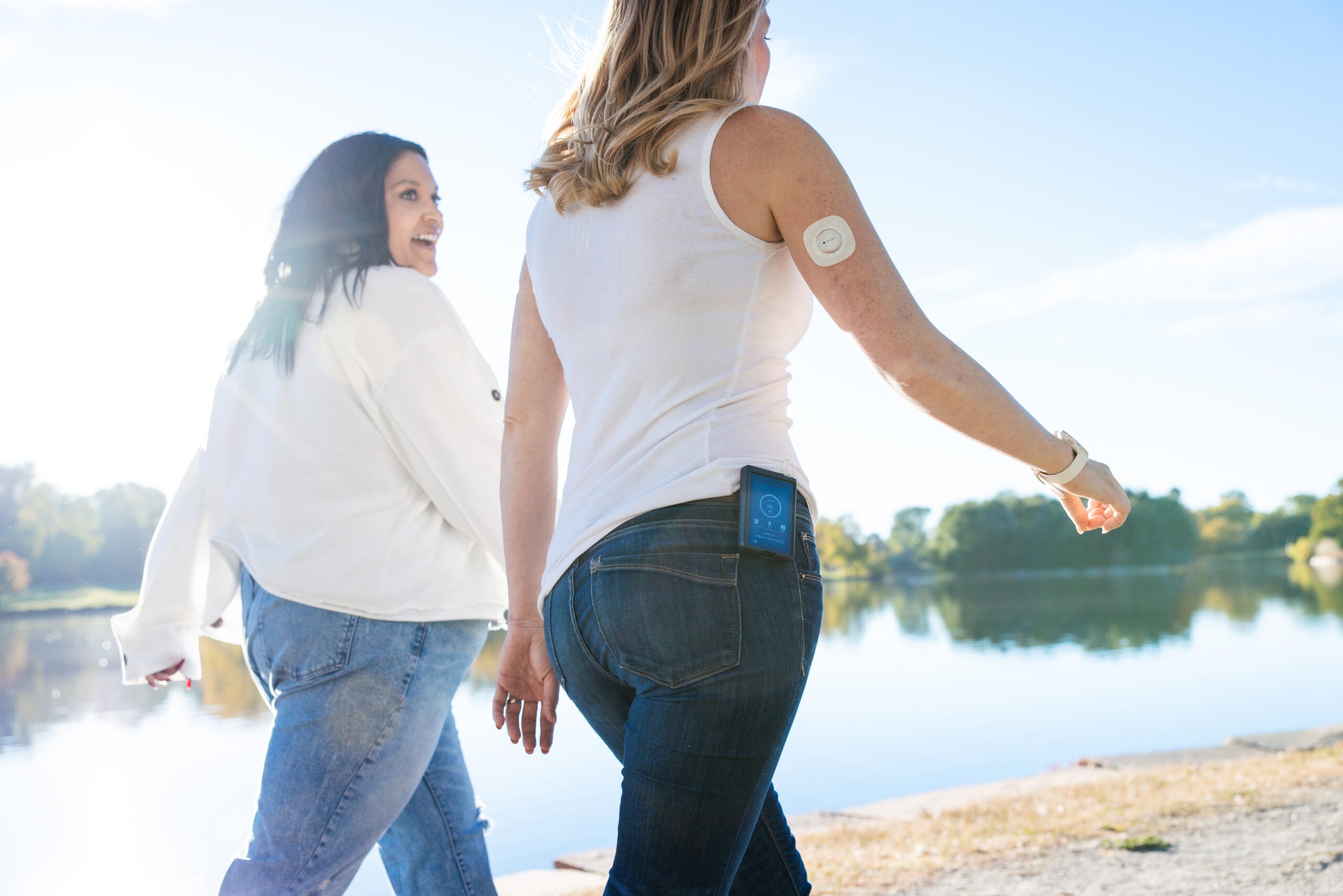 Two women walking outdoors near a lake on a sunny day, one wearing a white tank top with a medical device on her upper arm and a monitor clipped to her jeans.