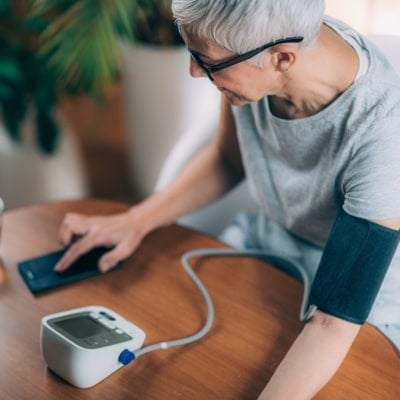 Senior woman wearing glasses and a gray shirt measuring her blood pressure with a digital monitor on a wooden table.