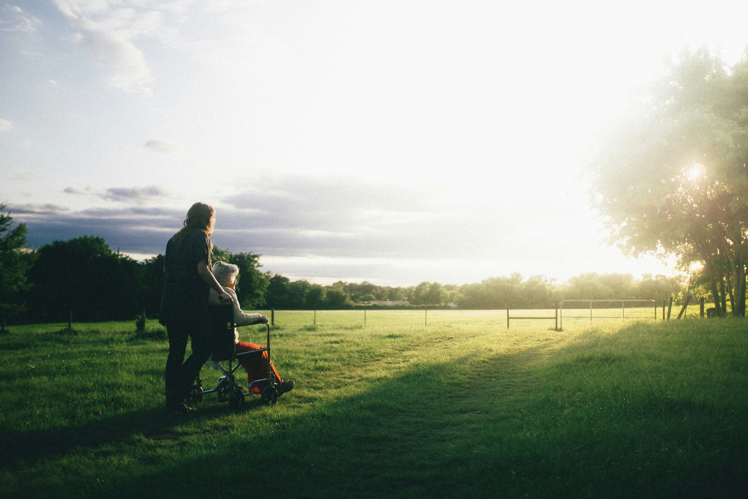 Person pushing an elderly woman in a wheelchair along a grassy field at sunset.