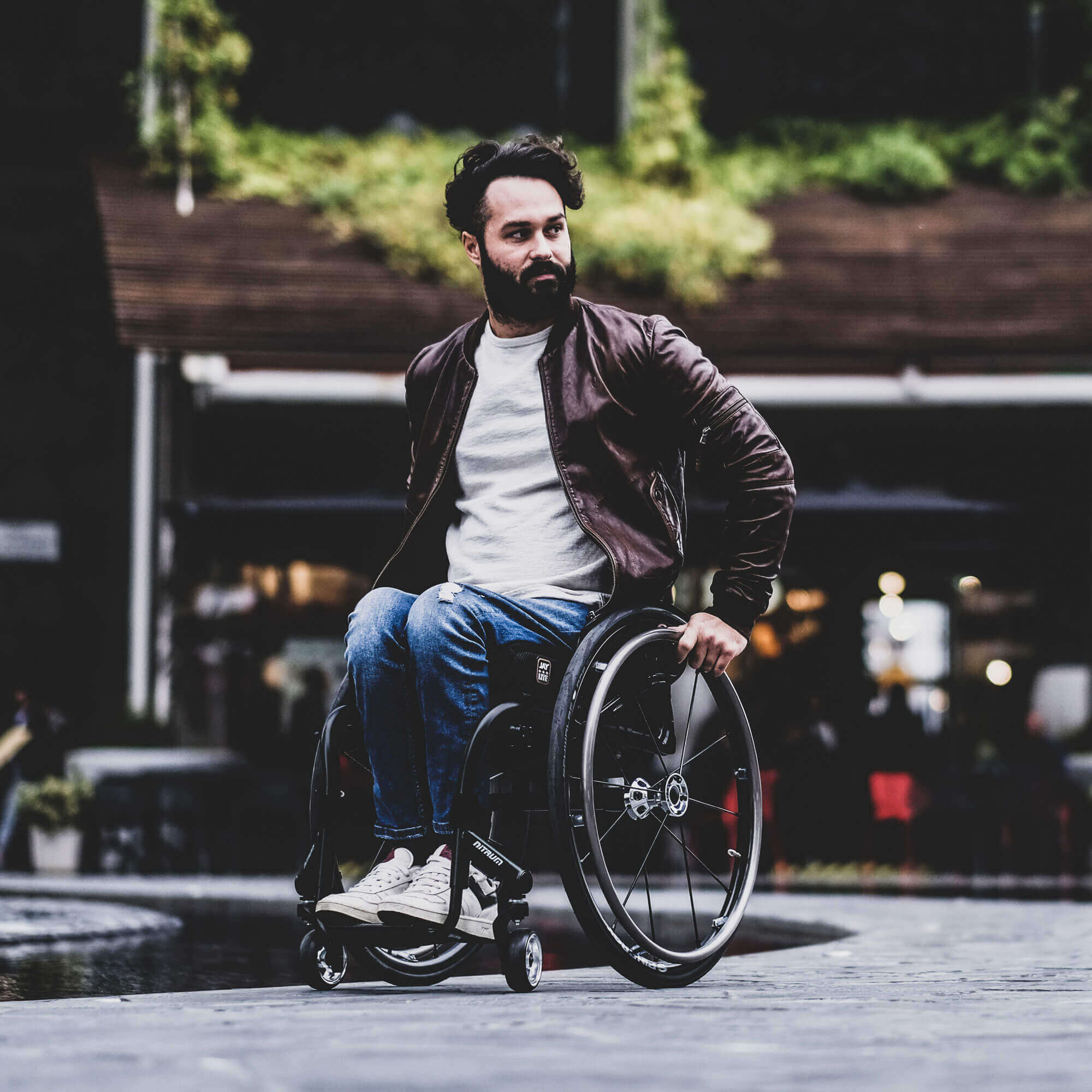 Man with a beard in a brown jacket and ripped jeans using a wheelchair outdoors on a paved surface.