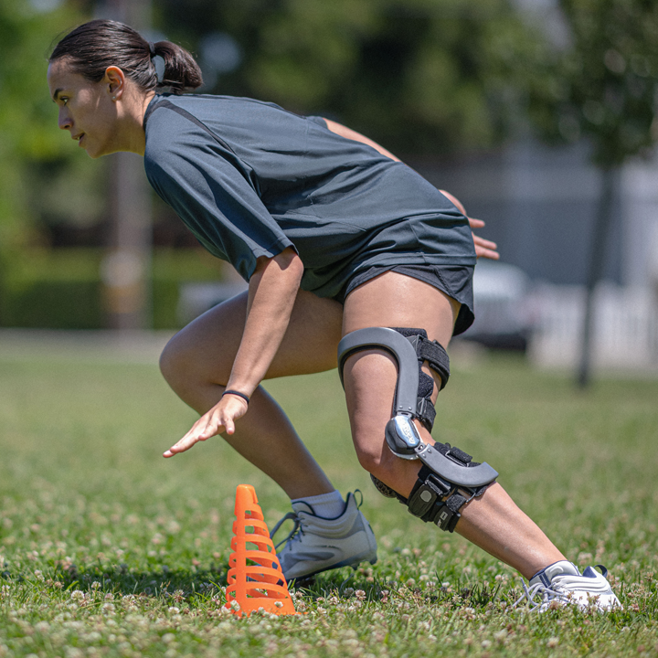 Athlete wearing a knee brace crouching and preparing to sprint next to an orange cone on grass.