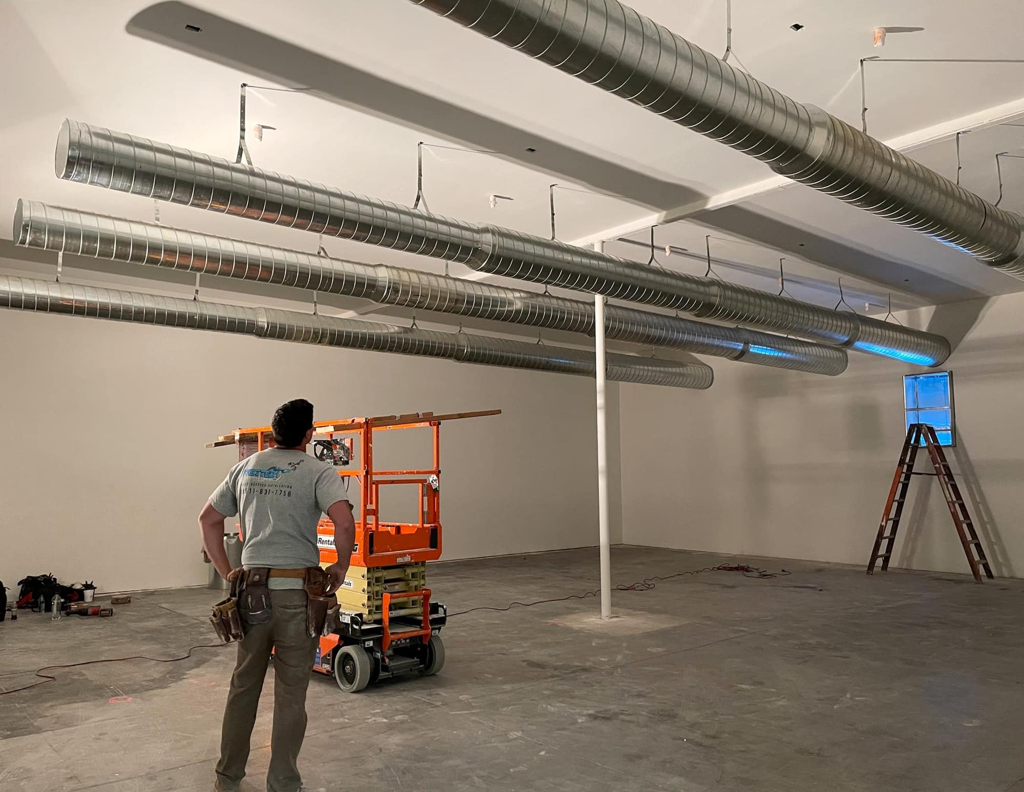 Construction worker with a tool belt standing in an unfinished room with exposed metal ductwork on the ceiling, an orange scissor lift, and a ladder near a window.