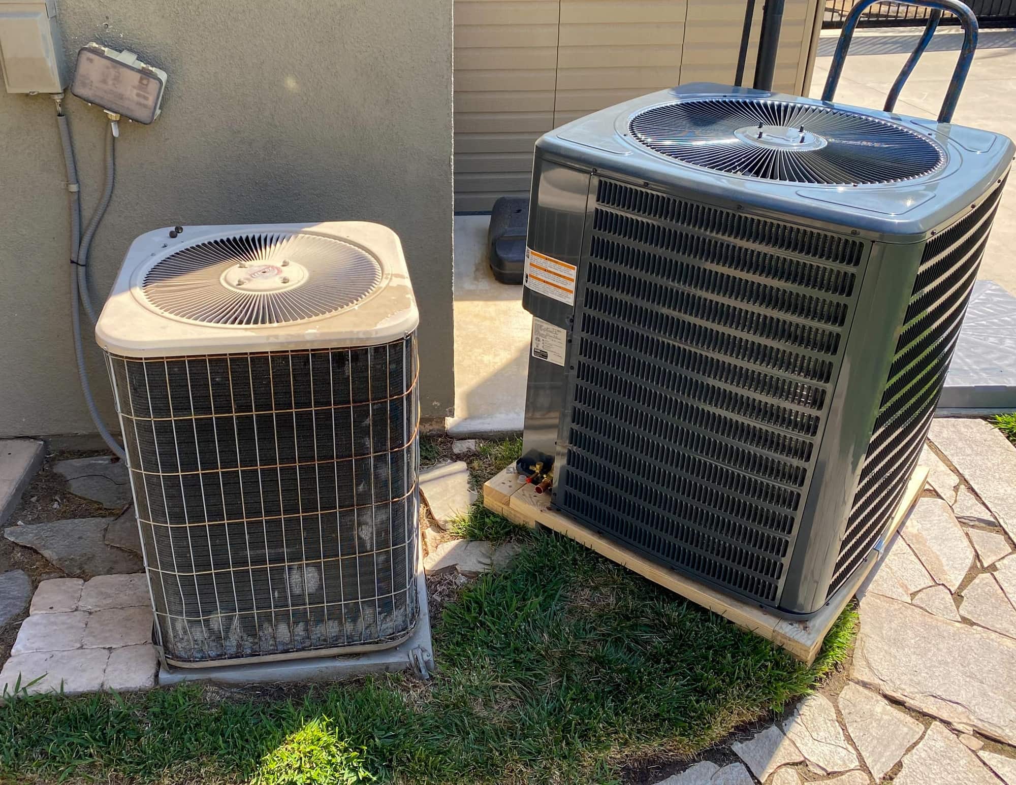 Two outdoor air conditioning units on concrete and grass near a building wall.