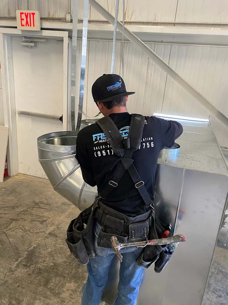 Worker wearing harness and tool belt assembling or inspecting metal HVAC ductwork inside an industrial building near an exit door.