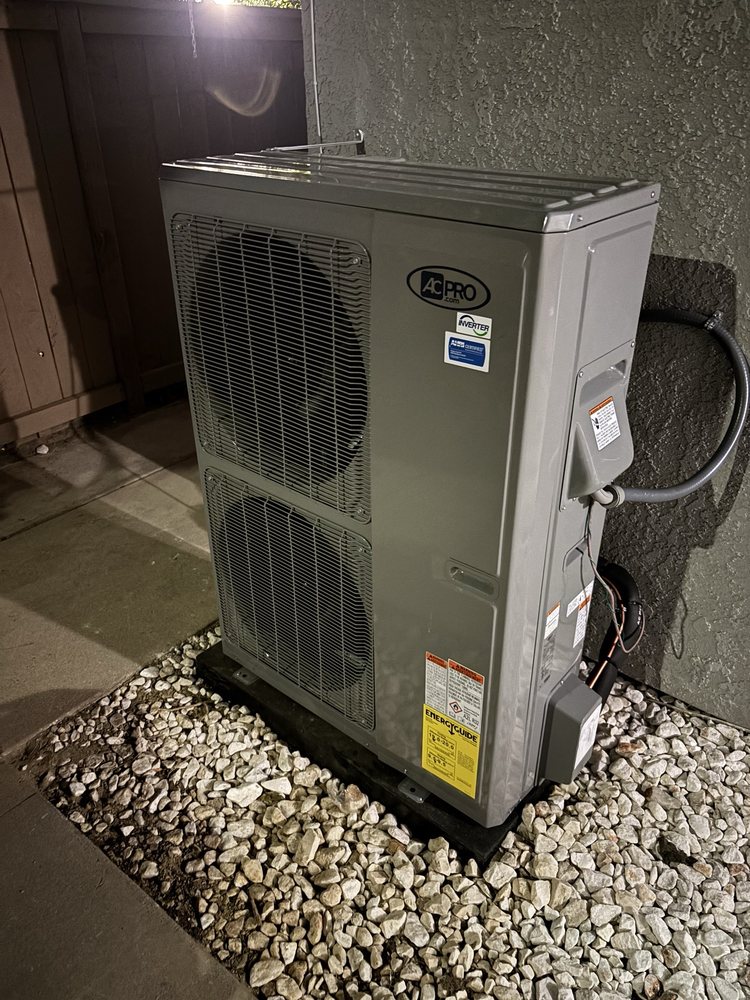 Outdoor heat pump unit installed on a black platform over white gravel beside a textured wall, captured at night.