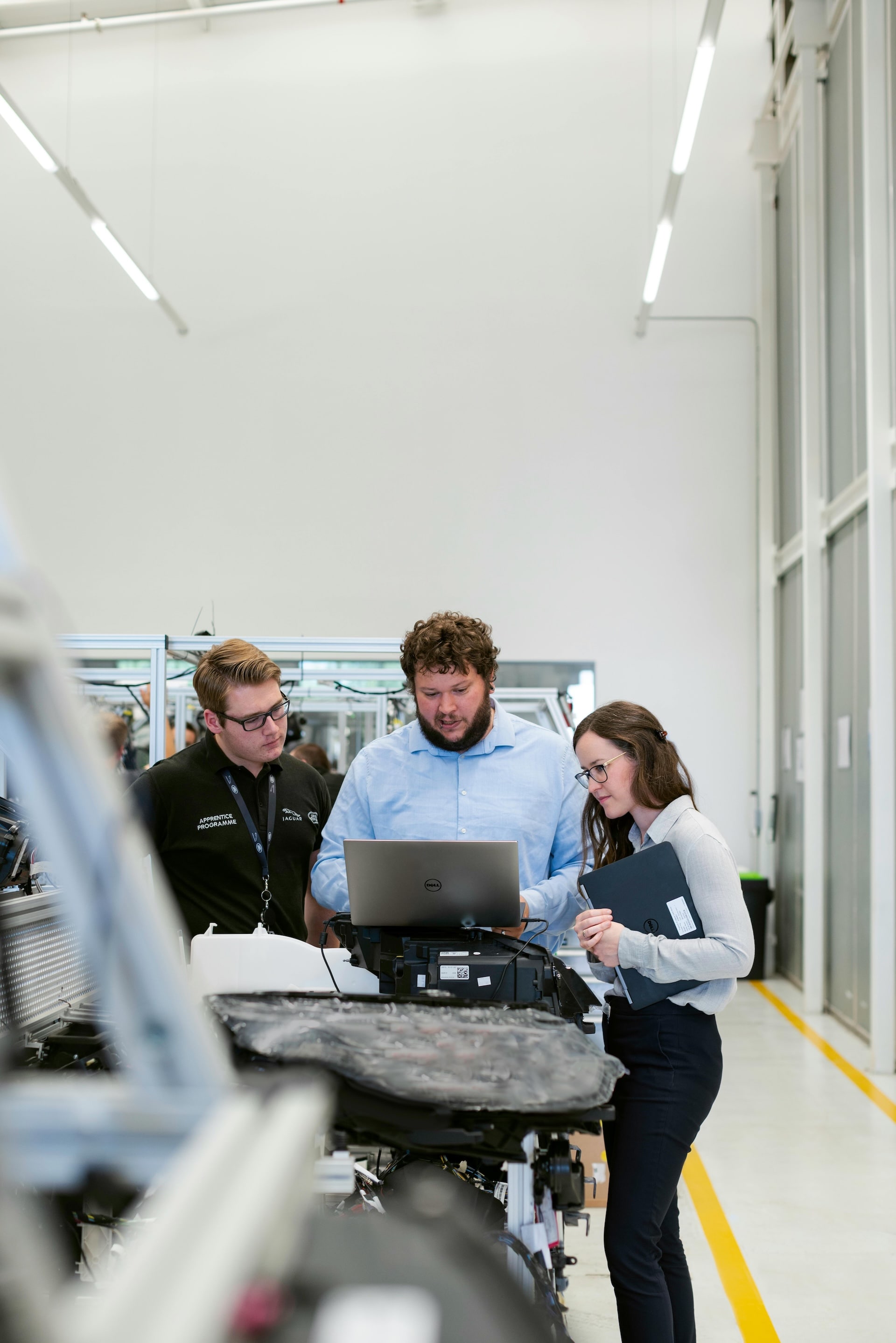 Three professionals in an industrial setting examine a laptop screen together near machinery.