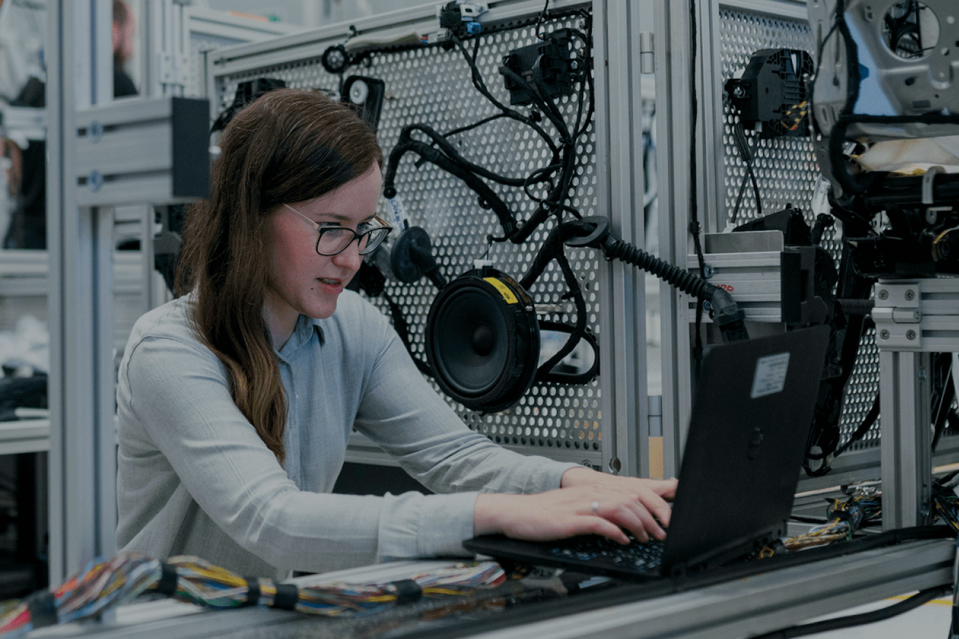 Woman wearing glasses working on a laptop in a technical workshop with electronic components and wiring around her.